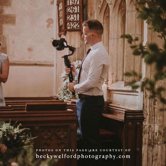 A photograph of Adam from Alchemist Designs in his role as wedding videographer. In this photo he is holding a camera and filming inside a church.
